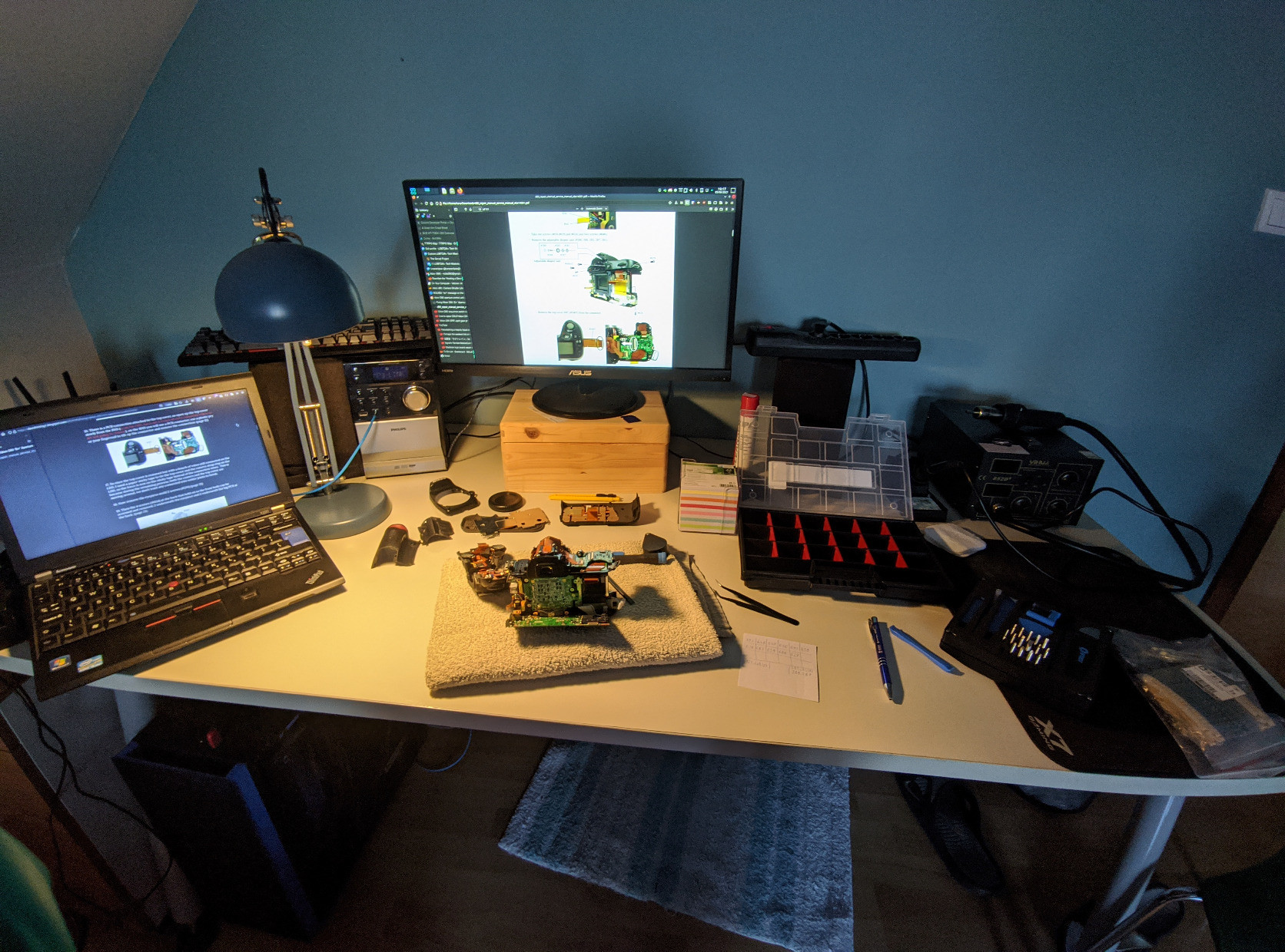 A photo showing my desk with some computer screens with the repair manual and a blog explaining the repair process. In the middle there's the parts shown in other pictures. To the right of those is a parts tray and some notes with screw numbers, among other tools.